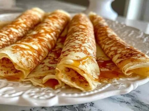Stack of golden oatmeal crepes on a white plate topped with fresh berries and a drizzle of maple syrup