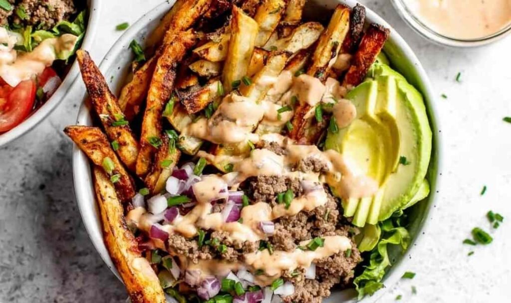 Paleo Burger Bowls with Fries topped with ground beef, crispy bacon, avocado, cherry tomatoes, pickles, and special sauce served in a white bowl