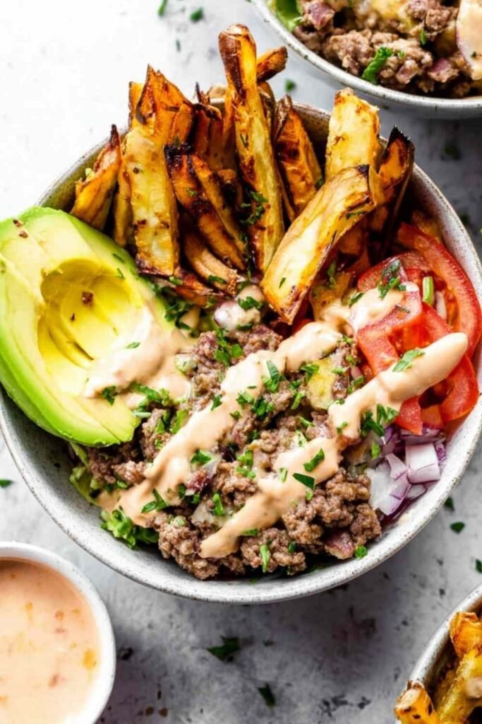 Paleo Burger Bowls with Fries topped with ground beef, crispy bacon, avocado, cherry tomatoes, pickles, and special sauce served in a white bowl