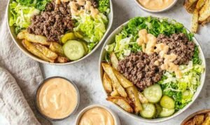 Overhead view of burger bowls with crispy potato chips, seasoned ground beef, shredded lettuce, cheese, pickles, and special sauce drizzle
