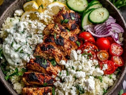 Easy Greek chicken bowls topped with sliced lemon-marinated chicken, feta, cucumber, tomatoes, and tzatziki sauce over rice