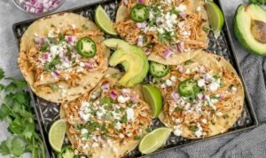 Healthy crockpot chicken tacos in flour tortillas with avocado, jalapenos, and lime on a wooden board