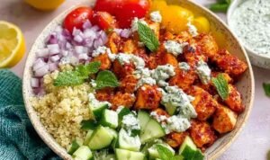 Four Mediterranean chicken bowls with harissa chicken, quinoa, tzatziki, and Greek salad on a wood table